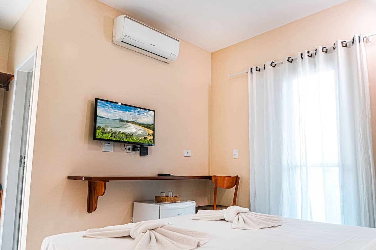 A hotel room with a bed, a television mounted on the wall, an air conditioning unit, a window with curtains, and a small table with a basket and bottles.