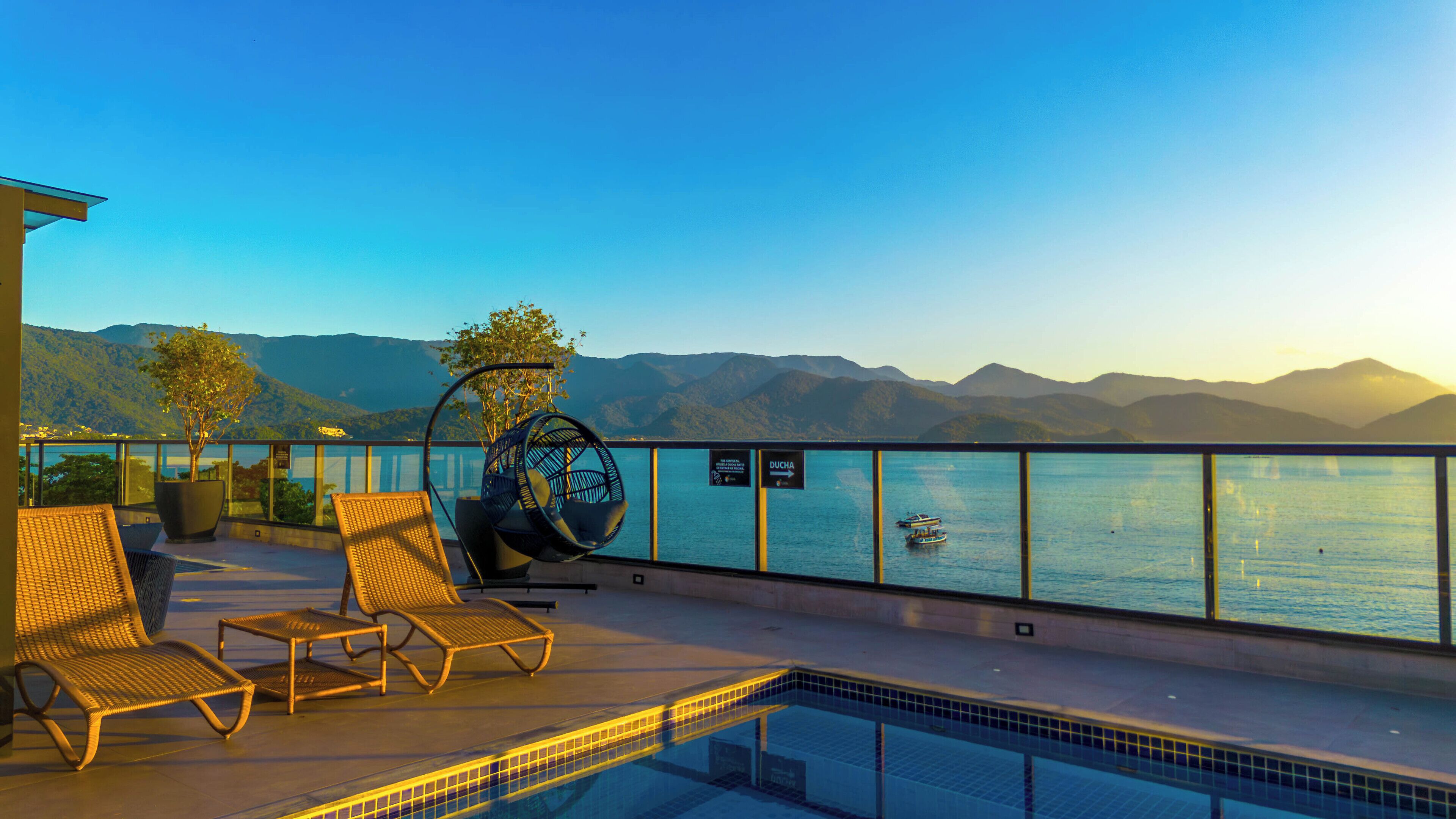 Zona de piscina con sillones de descanso y una hamaca colgante con vistas a un cuerpo de agua y montañas al fondo.