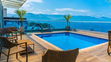 Zona de piscina con sillas de mimbre y plantas en macetas, con vista a un cuerpo de agua y montañas.