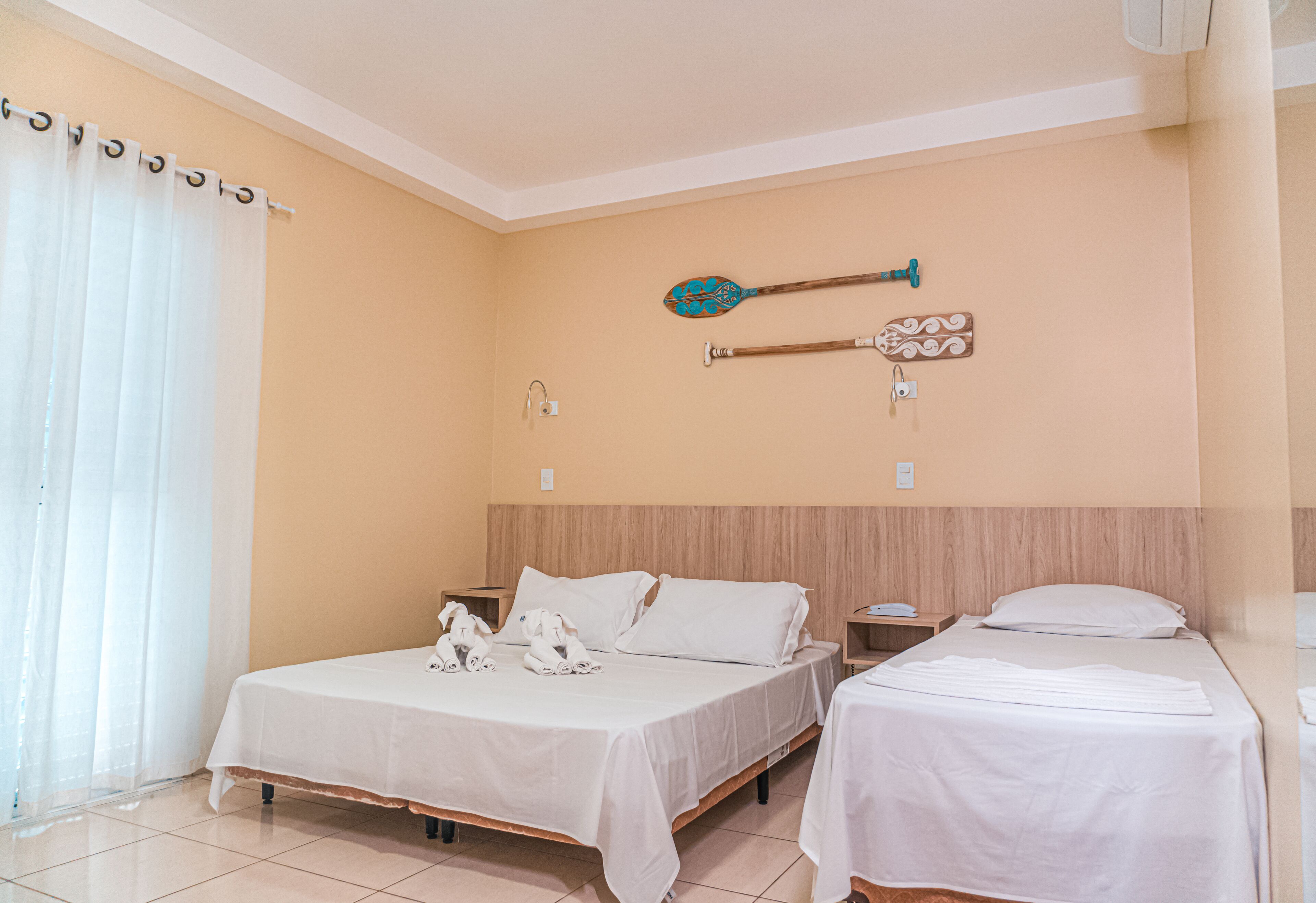 A hotel room with two beds, a wooden headboard, a wall-mounted paddle, and a window with sheer curtains.