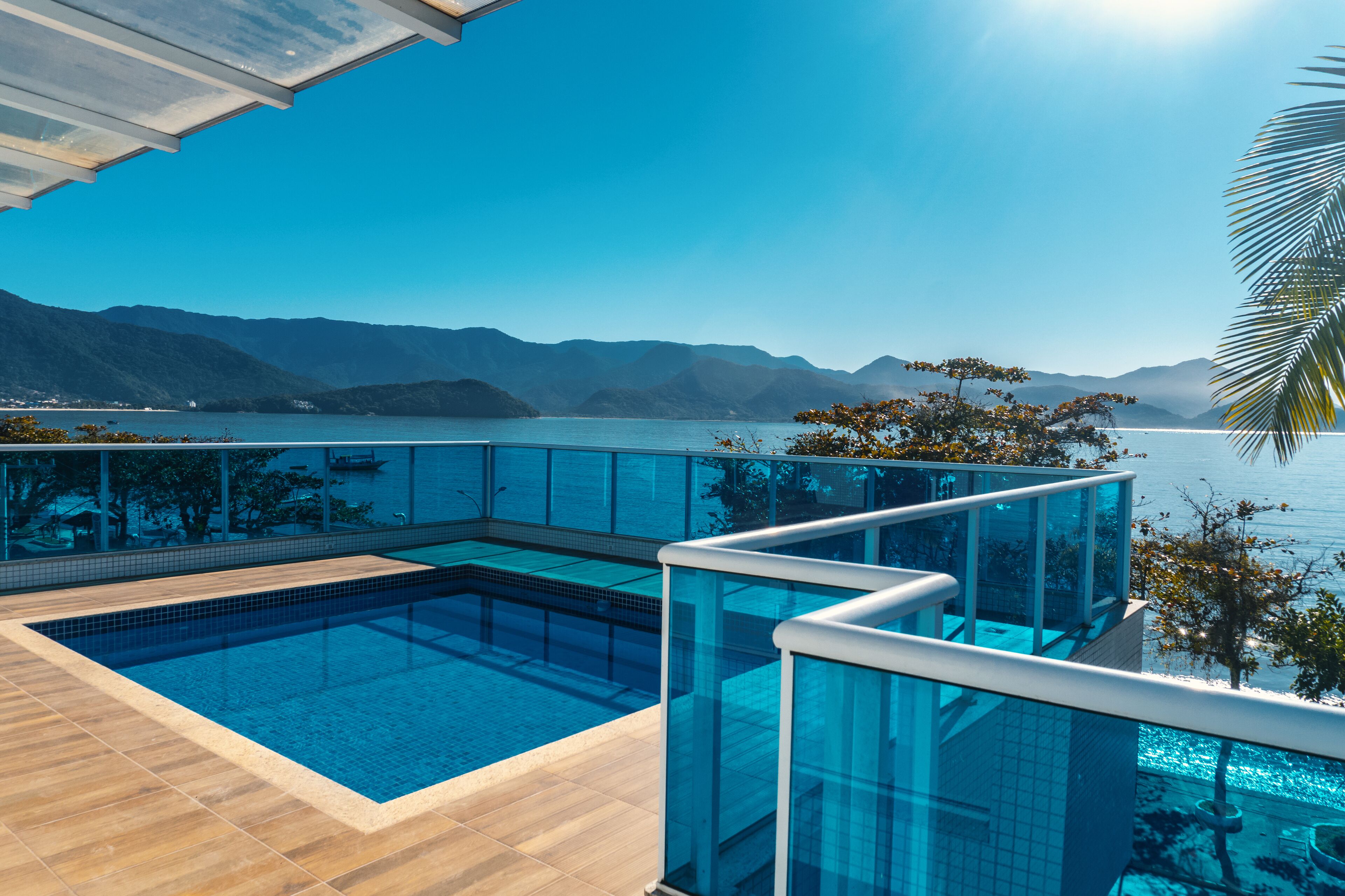 A modern infinity pool with clear blue water, surrounded by glass railings, overlooking a calm lake and distant mountains.