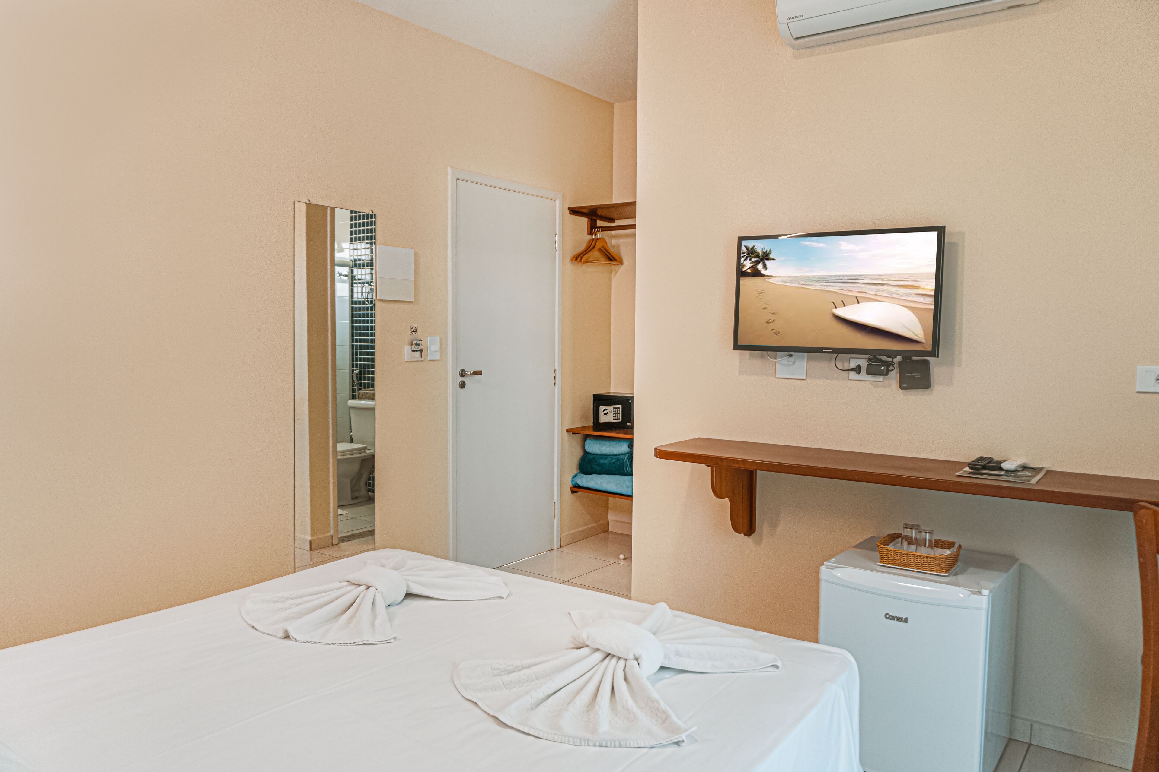 A hotel room with a bed, a small refrigerator, a television mounted on the wall, and a wooden shelf.