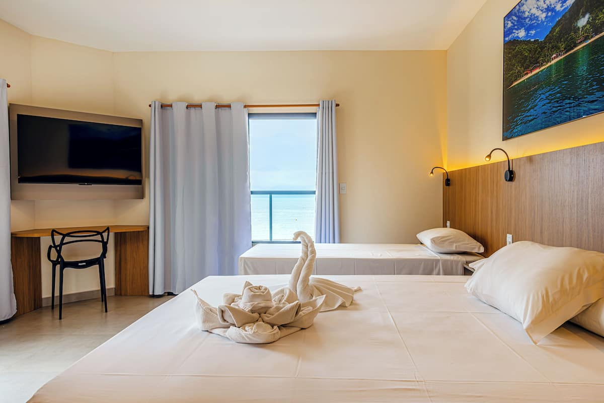 A hotel room with a bed, a TV, a desk with a chair, a window with curtains, and a wall-mounted light.