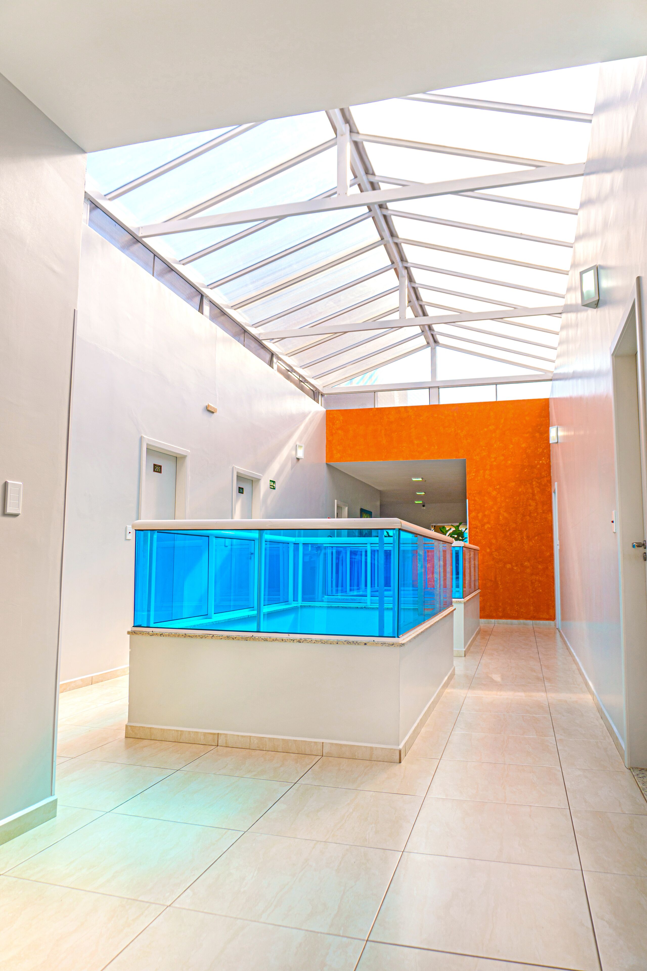 A modern hotel lobby with a glass ceiling, a central blue aquarium, and an orange accent wall.
