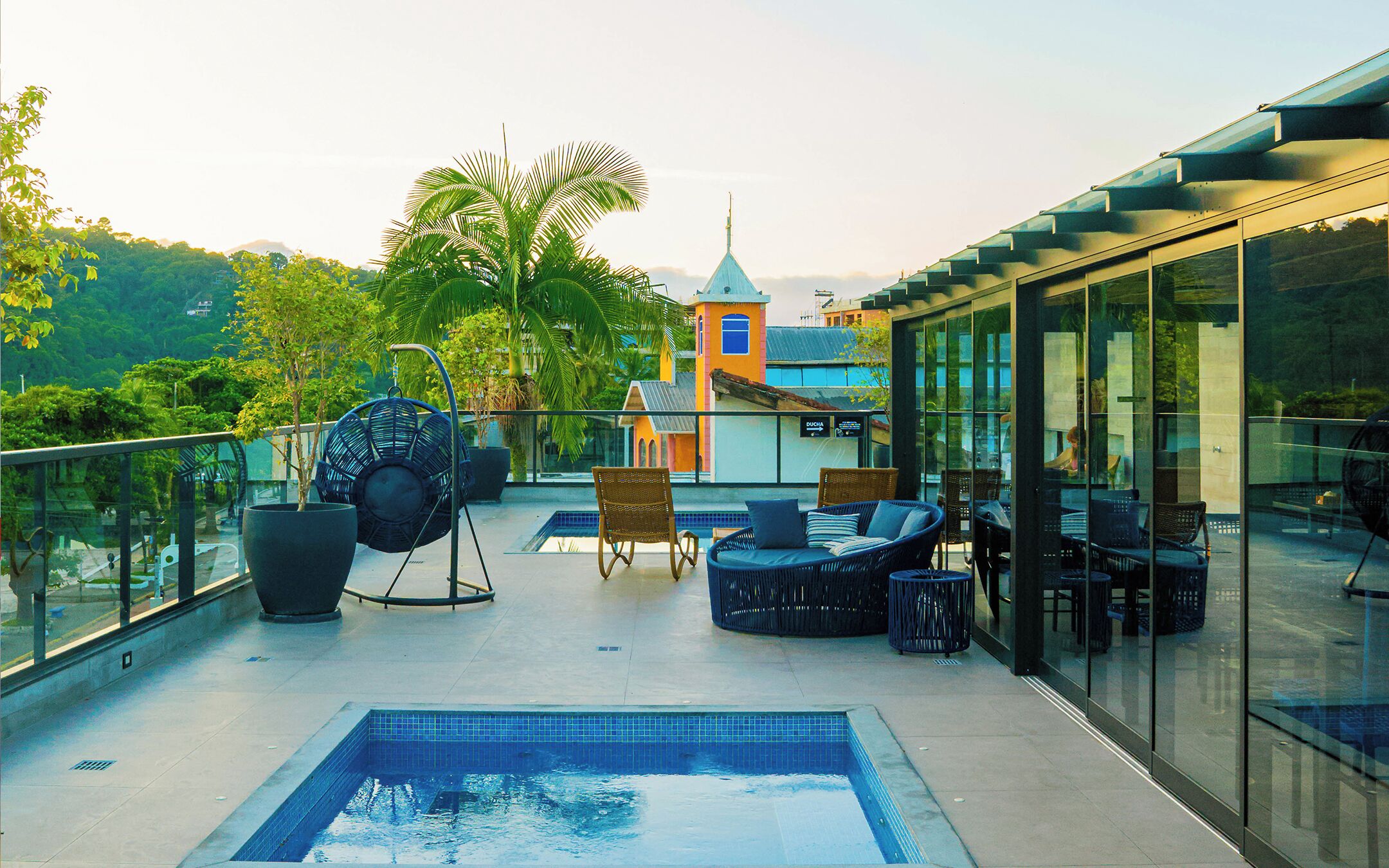 A rooftop pool area with a blue-tiled pool, wicker lounge chairs, and a glass-enclosed terrace with a view of a building and trees.