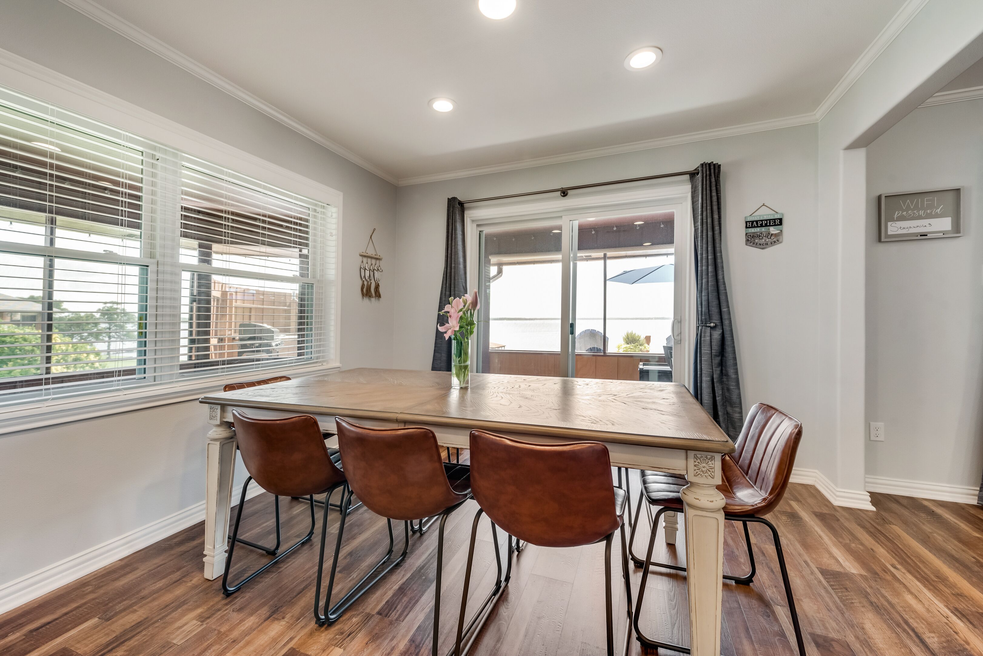 A dining area with a wooden table and four chairs, large windows with blinds, and a view of the outdoors.