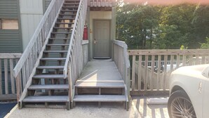 Eine Holztreppe führt zu einer Tür, in der Nähe steht ein weißes Auto und ein Holzzaun ist vorhanden.