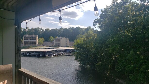Blick auf einen Fluss mit einem überdachten Anlegesteg, Gebäuden im Hintergrund und einer baumbewachsenen Uferpromenade.