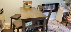 Un comedor con mesa y sillas de madera, un televisor montado en la pared y un estante con objetos decorativos.
