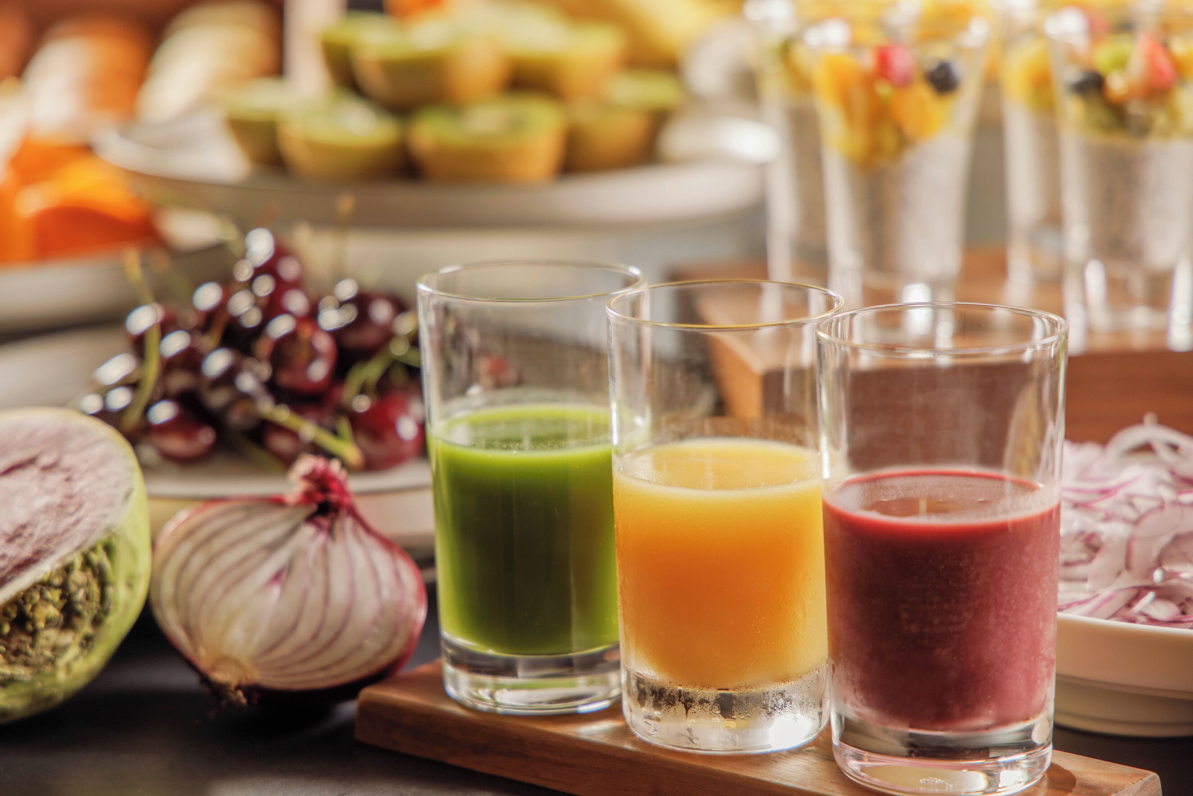 Tres vasos de jugo sobre una bandeja de madera, con un plato de cebollas en rodajas y un racimo de uvas al fondo.