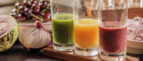 Tres vasos de jugo sobre una bandeja de madera, con un plato de cebollas en rodajas y un racimo de uvas al fondo.
