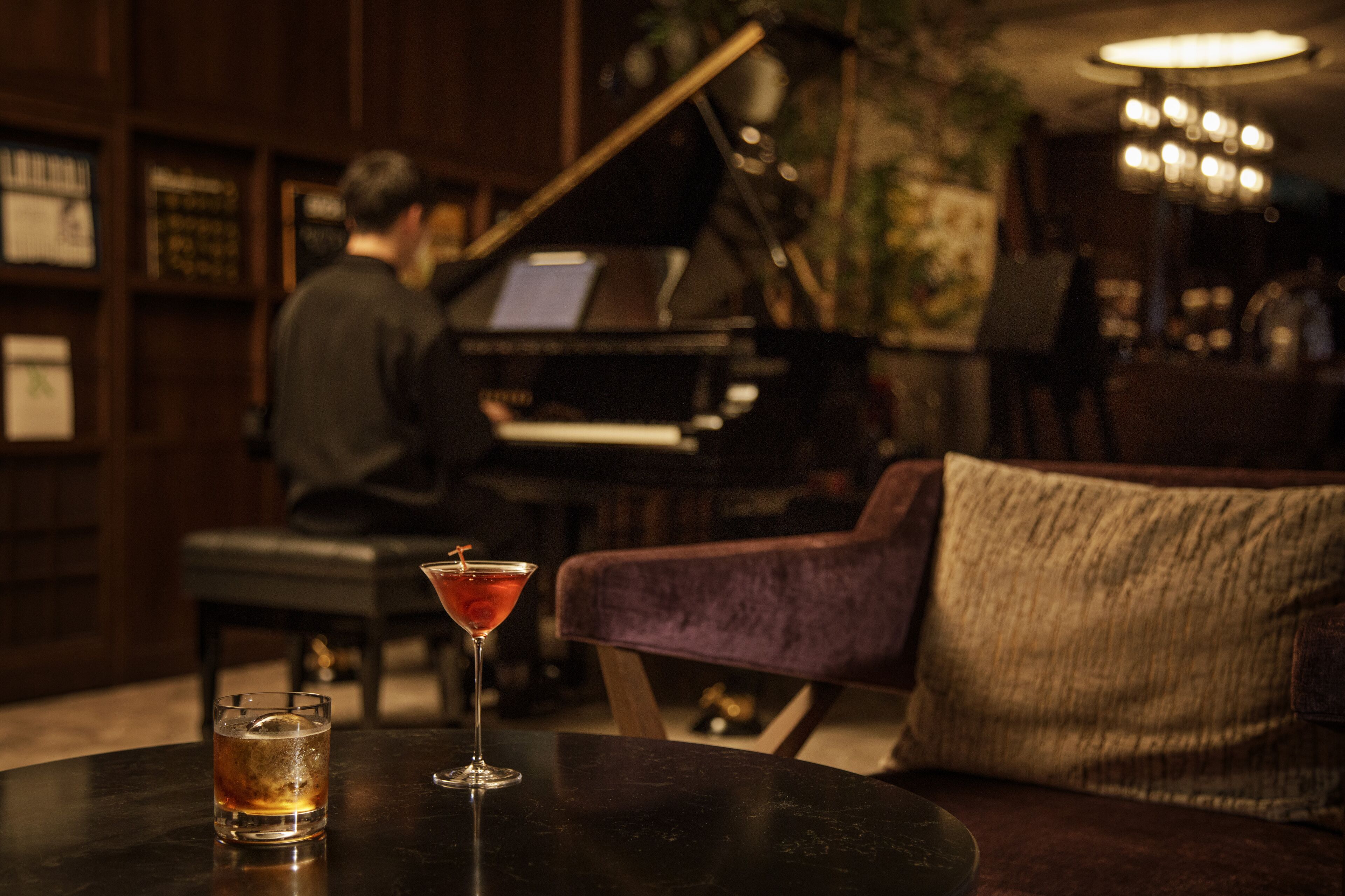 Zona de bar con un piano, una copa de martini y una copa de whiskey sobre una mesa.