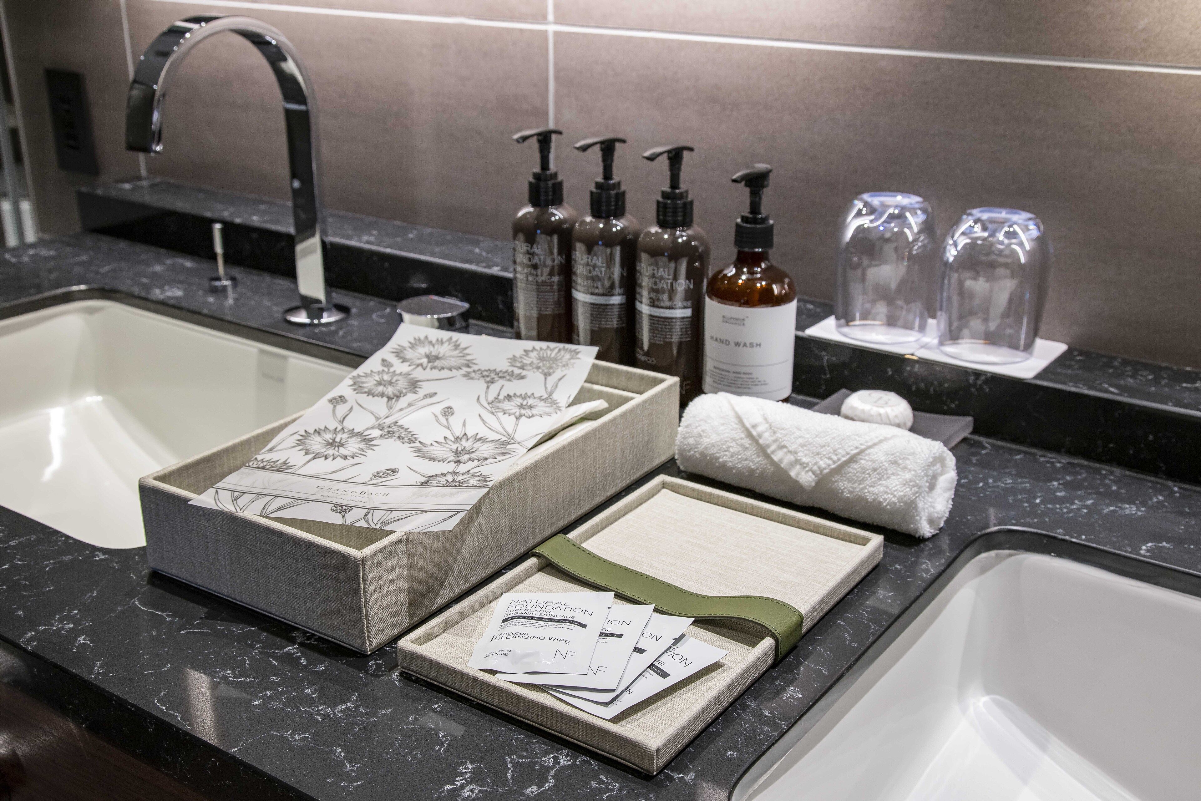 Un baño con encimera de mármol, lavamanos y espejo. Hay artículos de aseo personal, incluyendo botellas, una caja y una toalla.