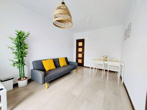 Una sala de estar moderna con un sofá gris, cojines amarillos, una lámpara colgante de fibras naturales, una mesa blanca con sillas y una alta planta de bambú.