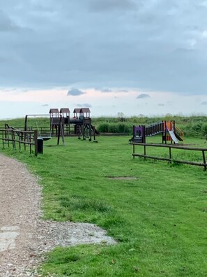 A playground with multiple play structures, including slides and climbing areas, surrounded by a wooden fence and grassy field.