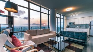 A modern living room with a sofa, armchairs, and a coffee table, large windows offering a city view, and a kitchen area with white cabinets and a countertop. - Spacious 3 Bedroom with stunning city view (Toronto)