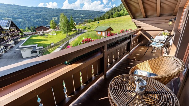 Ein Balkon mit Korbmöbeln, Blick auf eine grüne Landschaft und ein kleines Gebäude in der Ferne.