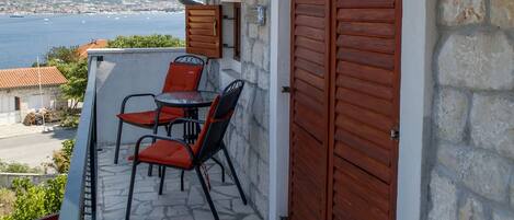 A balcony with a stone floor, a table with chairs, and a view of the sea and distant hills.