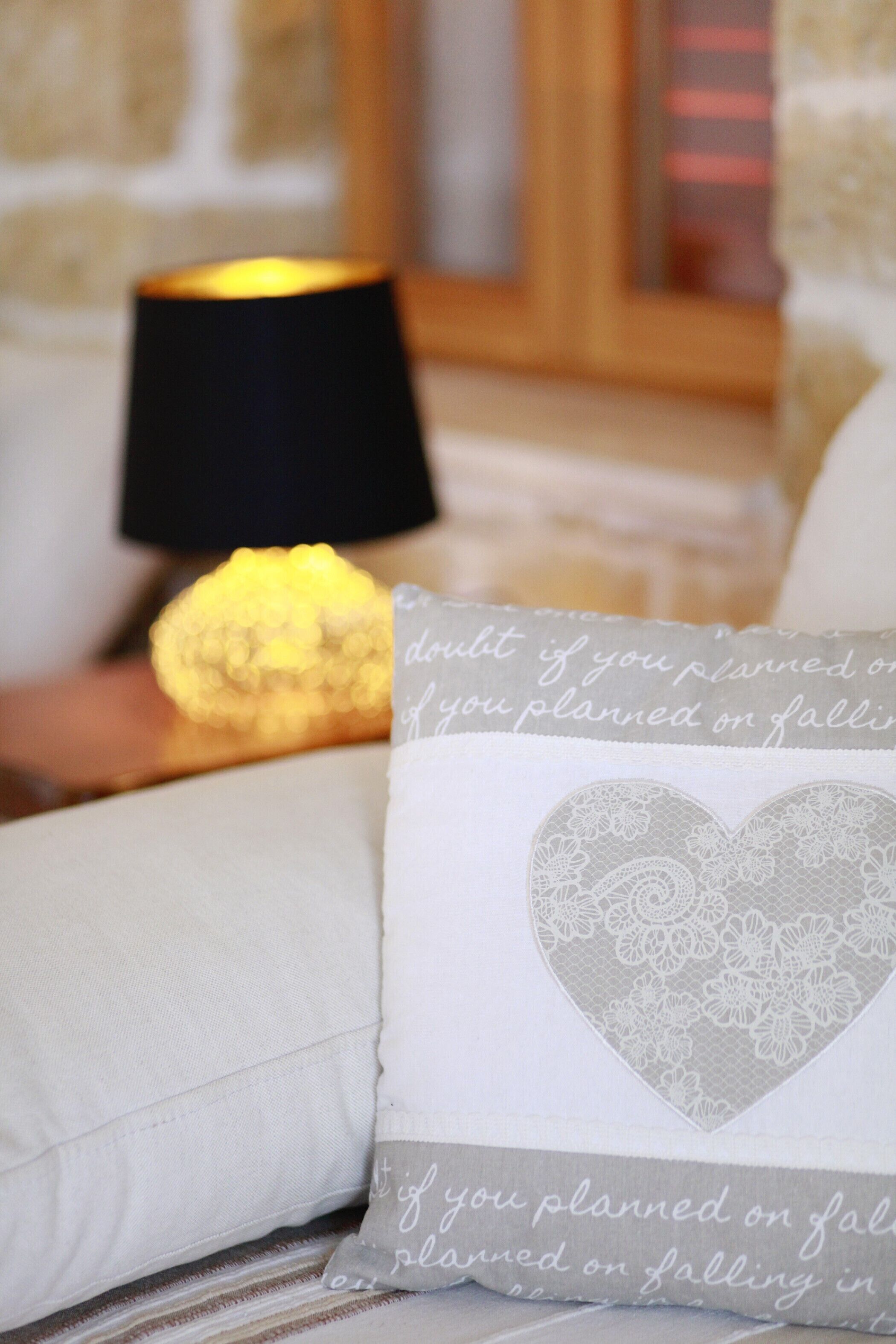 A white pillow with a lace heart design and a quote, a black lamp, and a yellow object on a white sofa.