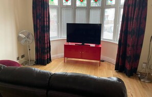 A living room with a flat-screen TV on a red cabinet, a black leather sofa, a standing fan, and a bay window with decorative glass panes. - Spacious 3BR Home in Bedford for Big Groups (Bedford)
