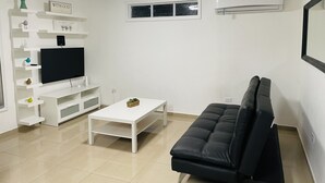 A living room with a flat-screen TV, white shelves, a black sofa, and a small window. - Hollywood Apartments (Yabucoa)