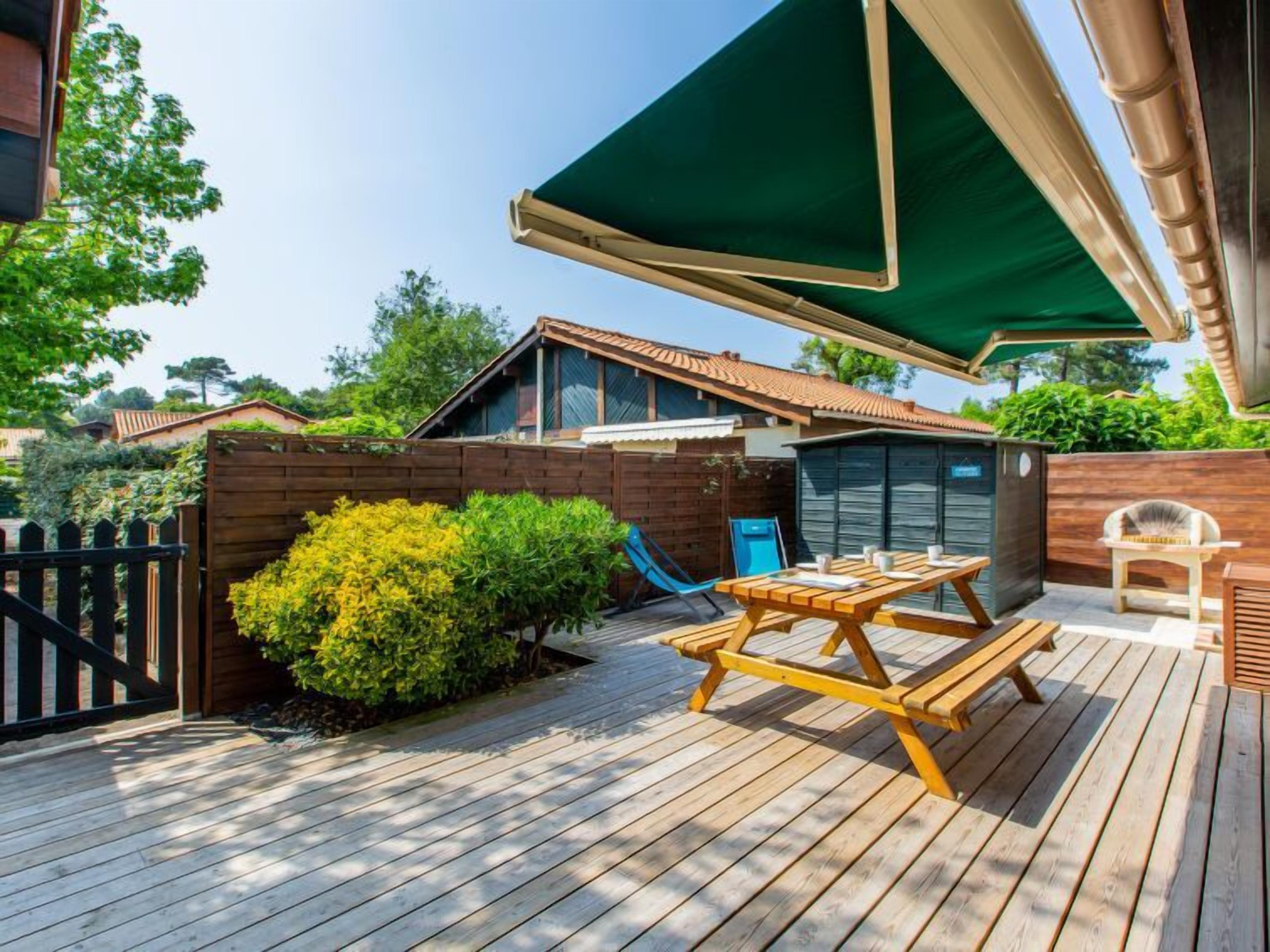 Una terraza de madera con una mesa y sillas de picnic, un paraguas verde y un cobertizo pequeño.