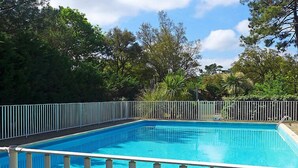 A rectangular swimming pool with a white metal fence surrounding it, set against a backdrop of lush green trees and a clear sky.