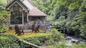 A cabin with a deck, surrounded by dense forest and a stream.