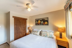 A bedroom with a bed, two bedside tables, a ceiling fan, a wall-mounted painting, and a door. - Snowdance 203c by Summit County Mountain Retreats (Keystone)