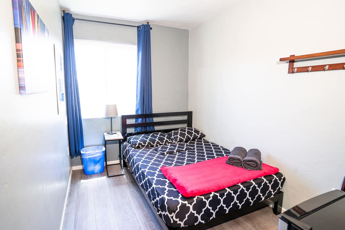Une chambre à coucher avec un lit, une table de chevet, une lampe, une poubelle bleue et une fenêtre avec des rideaux bleus.
