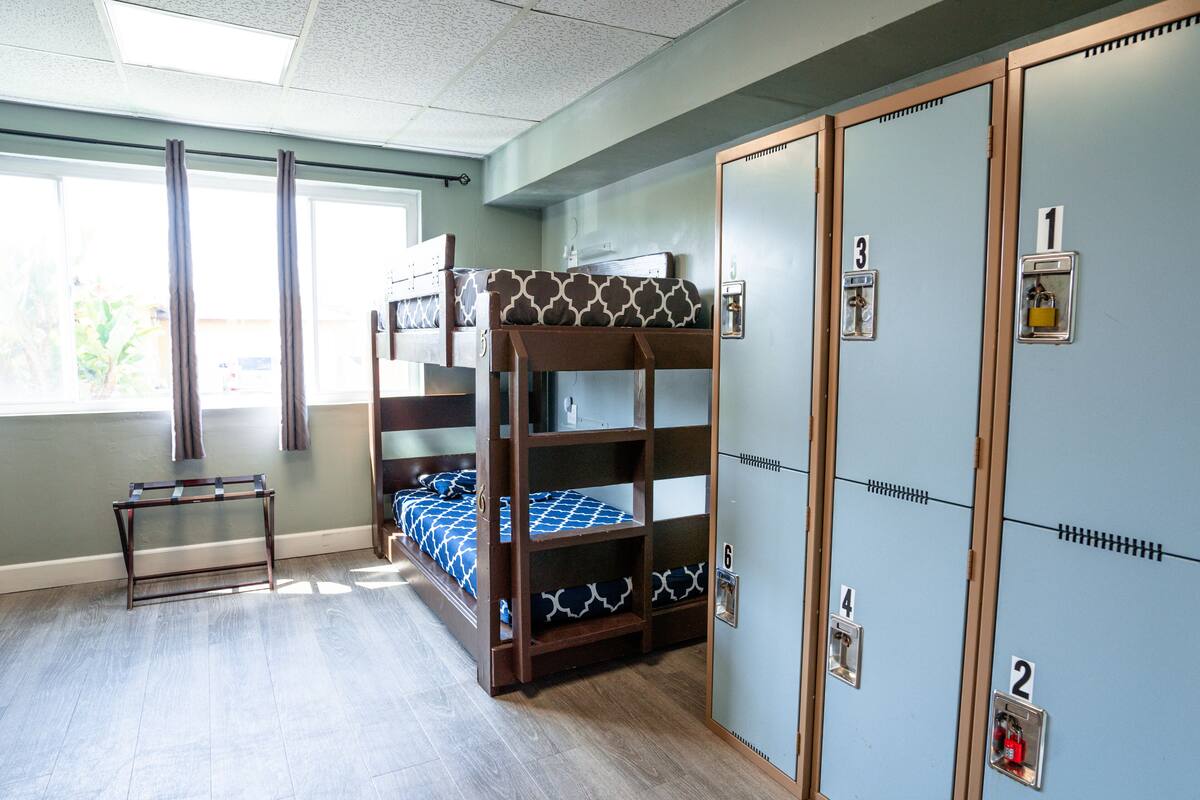 Une chambre avec un lit superposé, une petite table, une fenêtre avec des rideaux et un ensemble de casiers.
