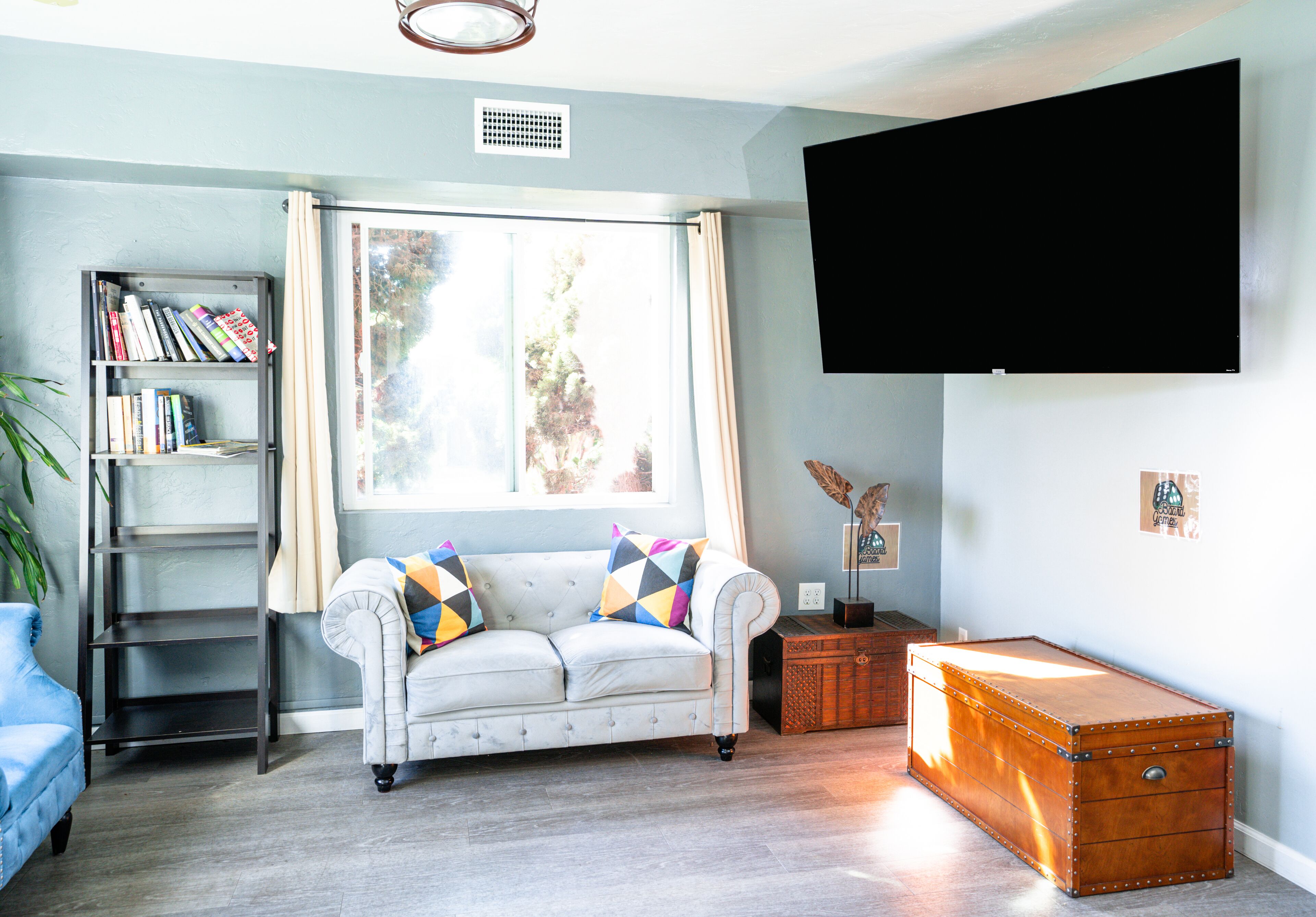 A living room with a white sofa, a bookshelf, a flat-screen TV, a window with curtains, and a vintage suitcase.