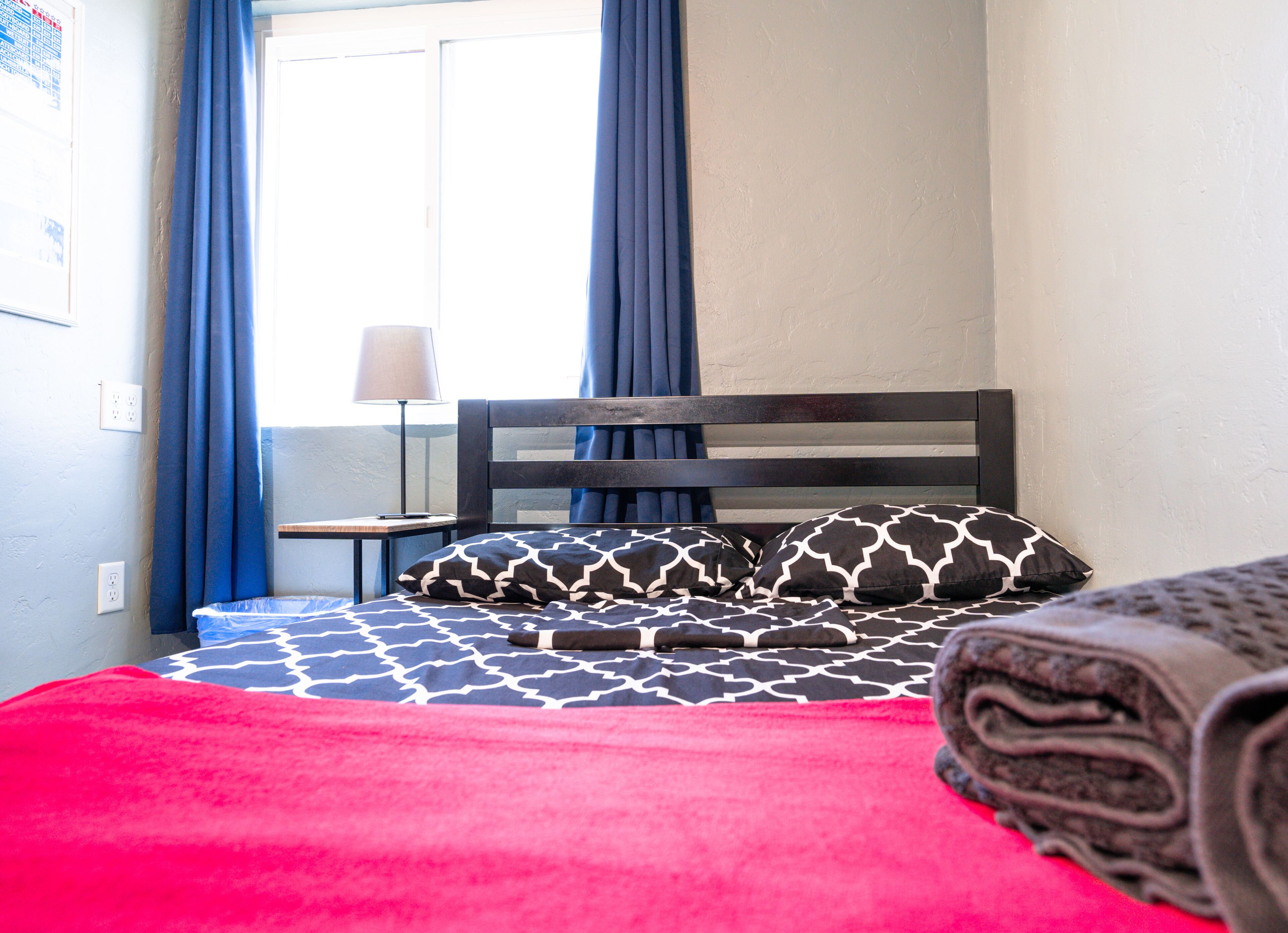 Una cama con ropa de cama estampada, una colcha roja y una manta enrollada en un asiento junto a la ventana.