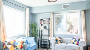 Una sala de estar con pared azul claro, un sofá blanco con cojines de colores, un sillón azul y una estantería.