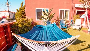 Una hamaca en un patio trasero con un edificio rojo, una silla azul y una tabla de surf.