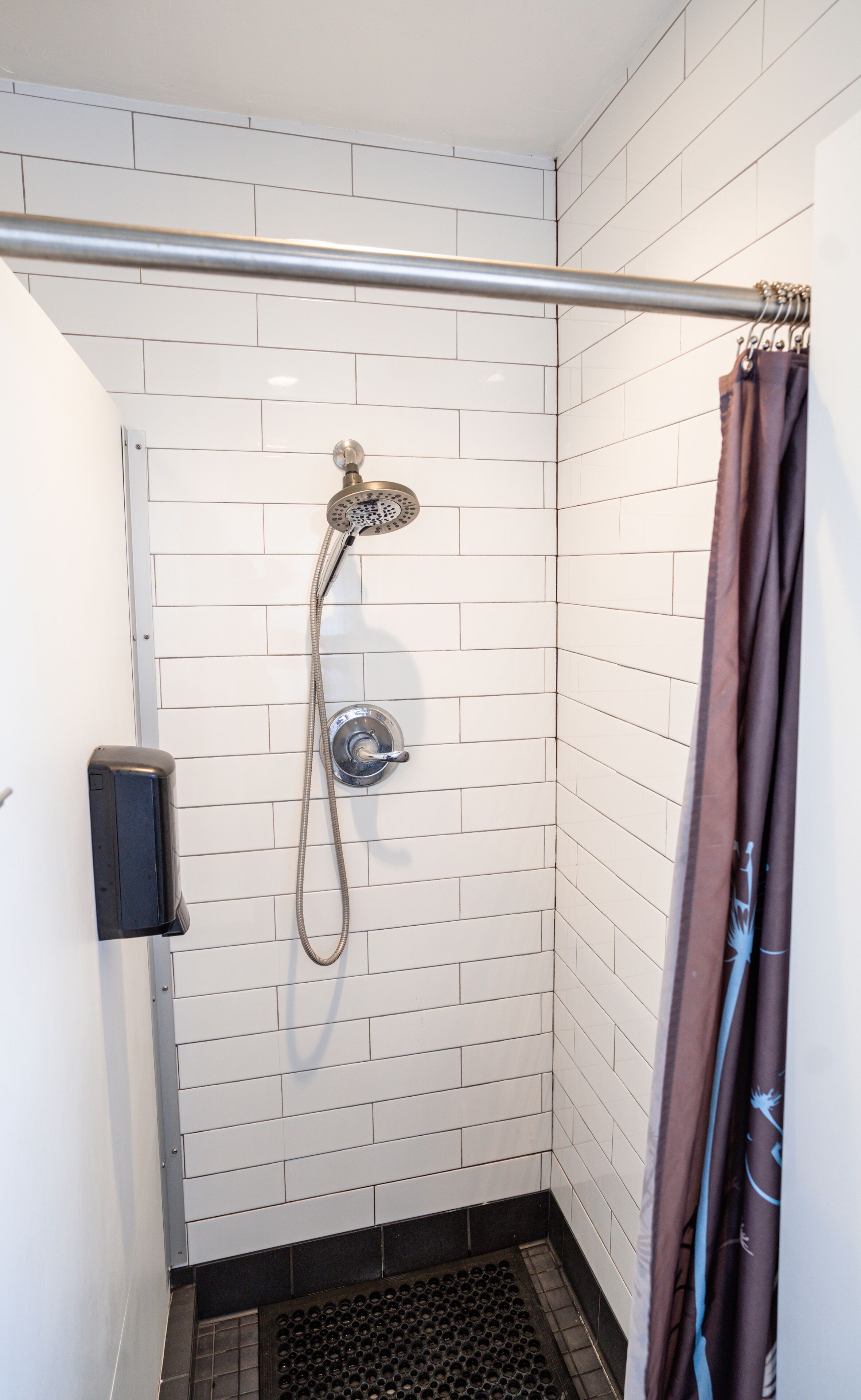 A shower area with white tiled walls, a showerhead, a curtain, and a drain.