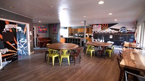 Un comedor amplio con varias mesas y sillas de madera, una cocina al fondo y un mural colorido en la pared.