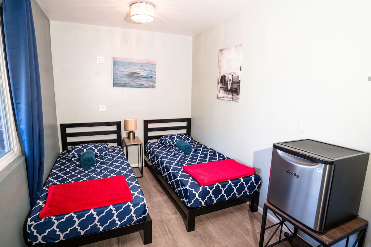 Une chambre avec deux lits, une petite table, un réfrigérateur et une fenêtre avec des rideaux.