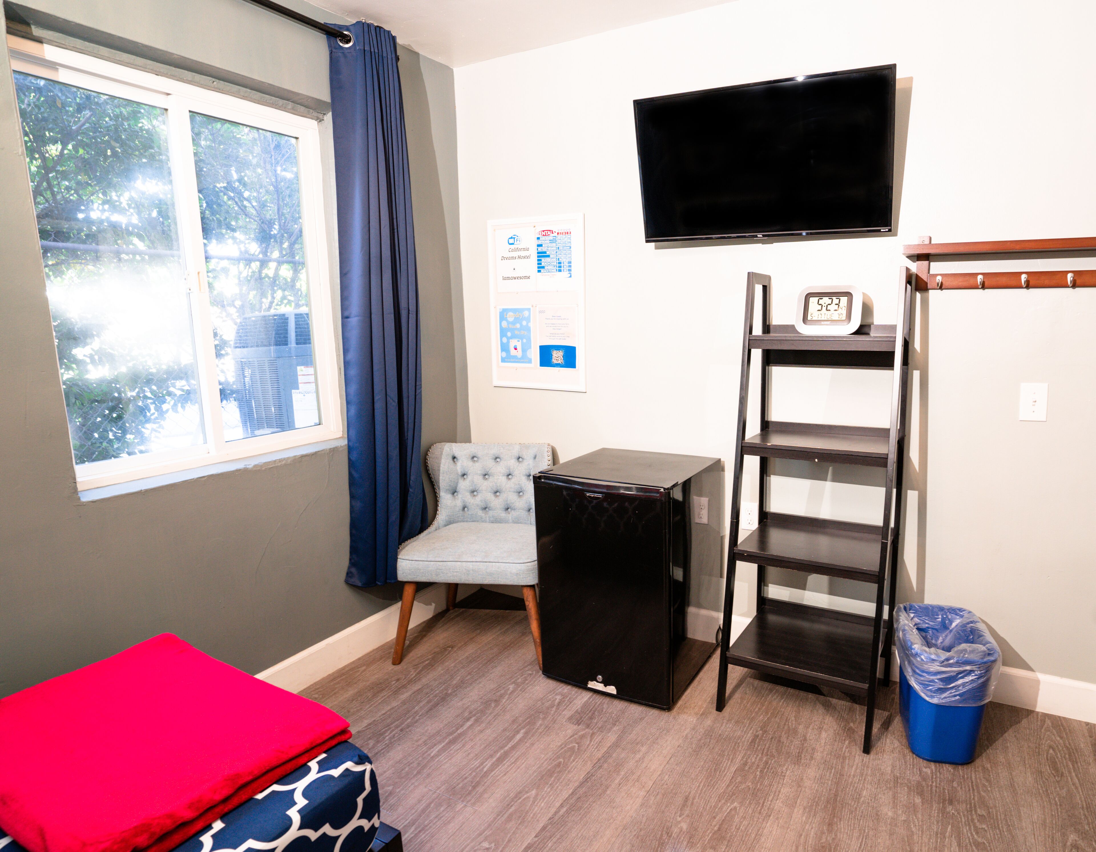 A small room with a bed, a chair, a TV, a shelf, and a trash bin.