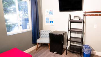 A small room with a bed, a chair, a TV, a shelf, and a trash bin.