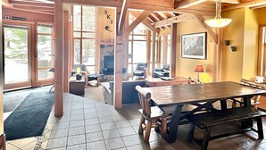 A spacious room with a wooden ceiling, large windows, a fireplace, and a dining area with a table and chairs. - Large Ski-In / Ski-Out Timber Frame House, Open Concept, New Hot Tub! (Golden)