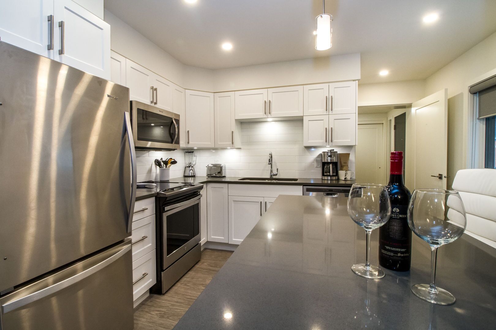 A modern kitchen with stainless steel appliances, white cabinetry, and a central island with wine glasses and a bottle.