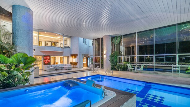 Una piscina interior con cubierta de madera, un área de descanso con asientos y ventanas amplias que ofrecen vistas del paisaje urbano.