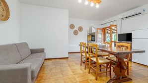 A living room with a sofa, dining table with chairs, a TV, and a kitchen area with an air conditioner.