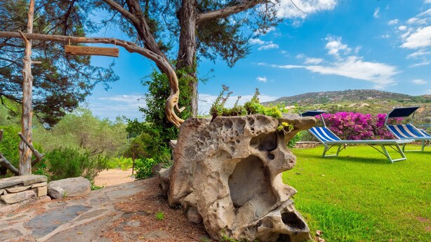 A stone structure with a carved hole, a wooden sign, and two blue and white striped lounge chairs in a grassy area with trees and a distant hill.