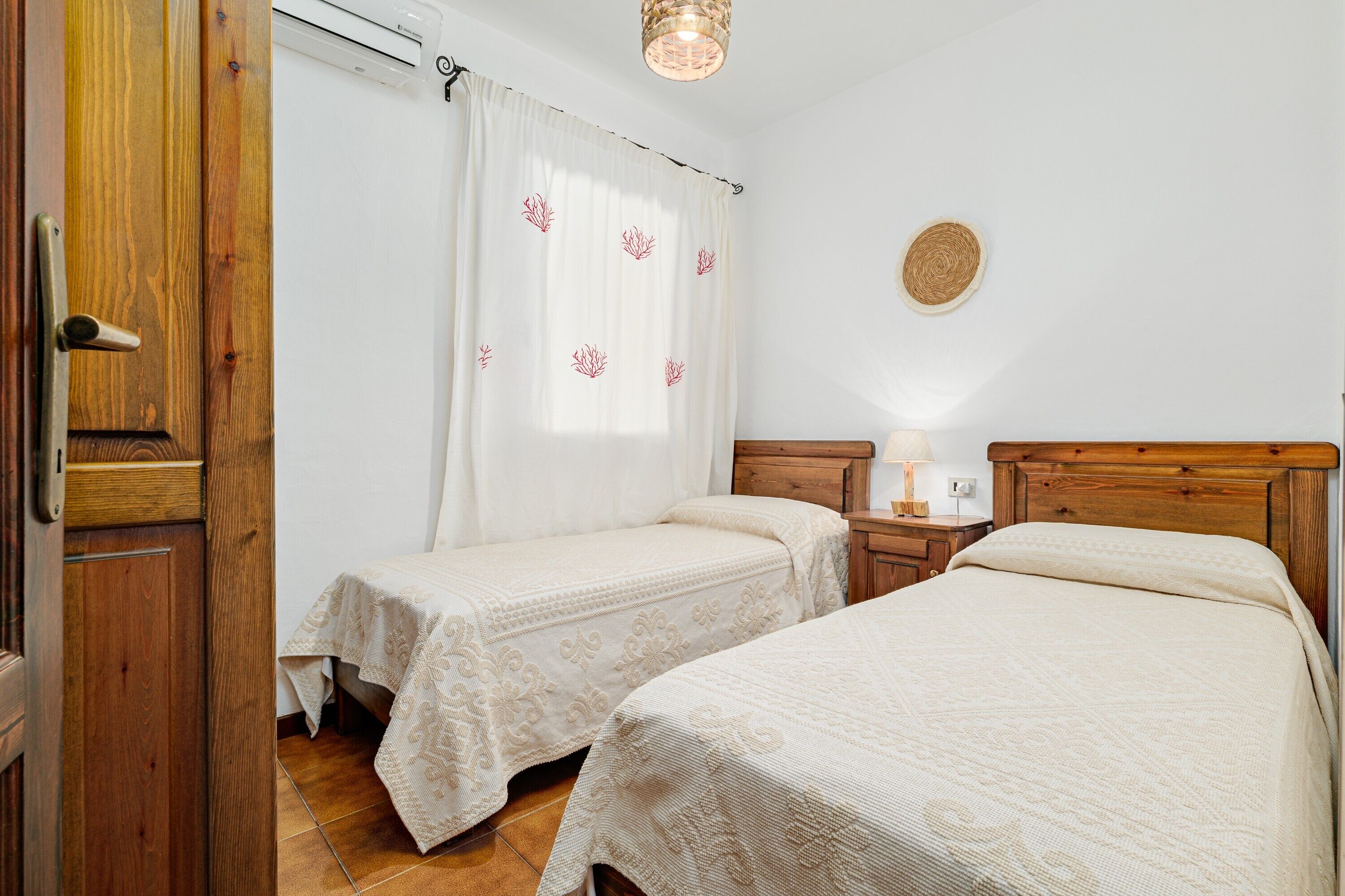 Une chambre avec deux lits, une porte en bois, un rideau blanc orné de motifs floraux, une table de chevet avec une lampe et un élément décoratif rond fixé au mur.
