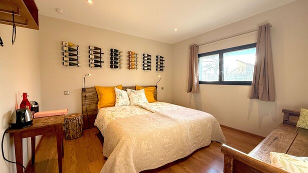 A bedroom with a large bed, wooden flooring, a bedside table with a lamp, a wall-mounted wine rack, and a window with curtains. - Chambre D'hôtes sur Domaine Viticole Proche Colmar Eguisheim sur Route des Vins (GUEBERSCHWIHR)