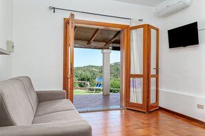 Una habitación con sofá, una puerta abierta que da a un balcón con una silla y un televisor en la pared.