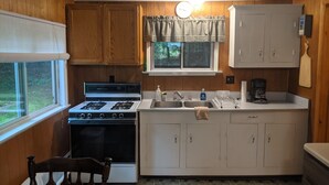 A compact kitchen with wooden cabinets, a stove, a sink, and a window with a view of greenery. - Moreno's Cottages Cabin #10 -2 Bed/2 Bath Adirondack 4 season Cabin in the pines (Saranac Lake)