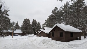 A snow-covered cabin area with multiple wooden cabins, a dirt path, and surrounding trees. - Moreno's Cottages Cabin #10 -2 Bed/2 Bath Adirondack 4 season Cabin in the pines (Saranac Lake)